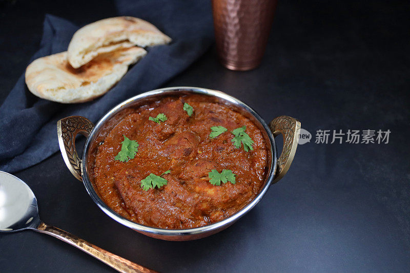 卡扎伊(kadhai)烹饪锅/上菜碗，里面盛满了用绿色香菜叶装饰的自制鸡肉布娜(bhuna)，配上印度烤饼，金属勺，黑色材料背景，聚焦前景，从高处看